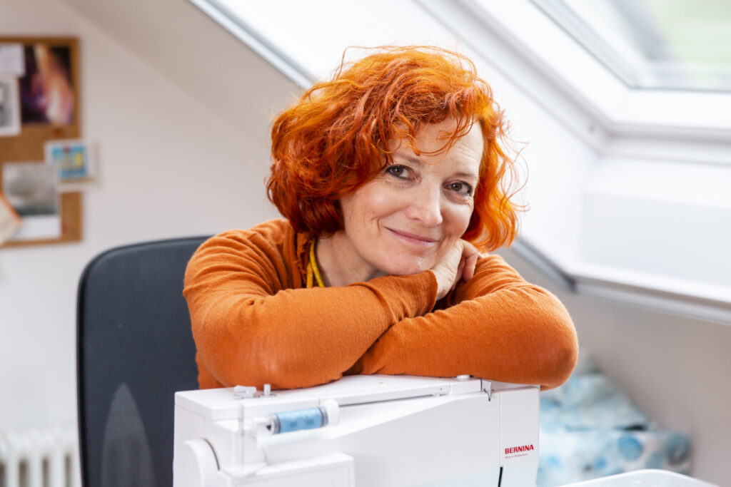 Porträt einer lächelnden Frau (Isabelle Wiesele) mit roten Haaren, an einen Tisch gelehnt.