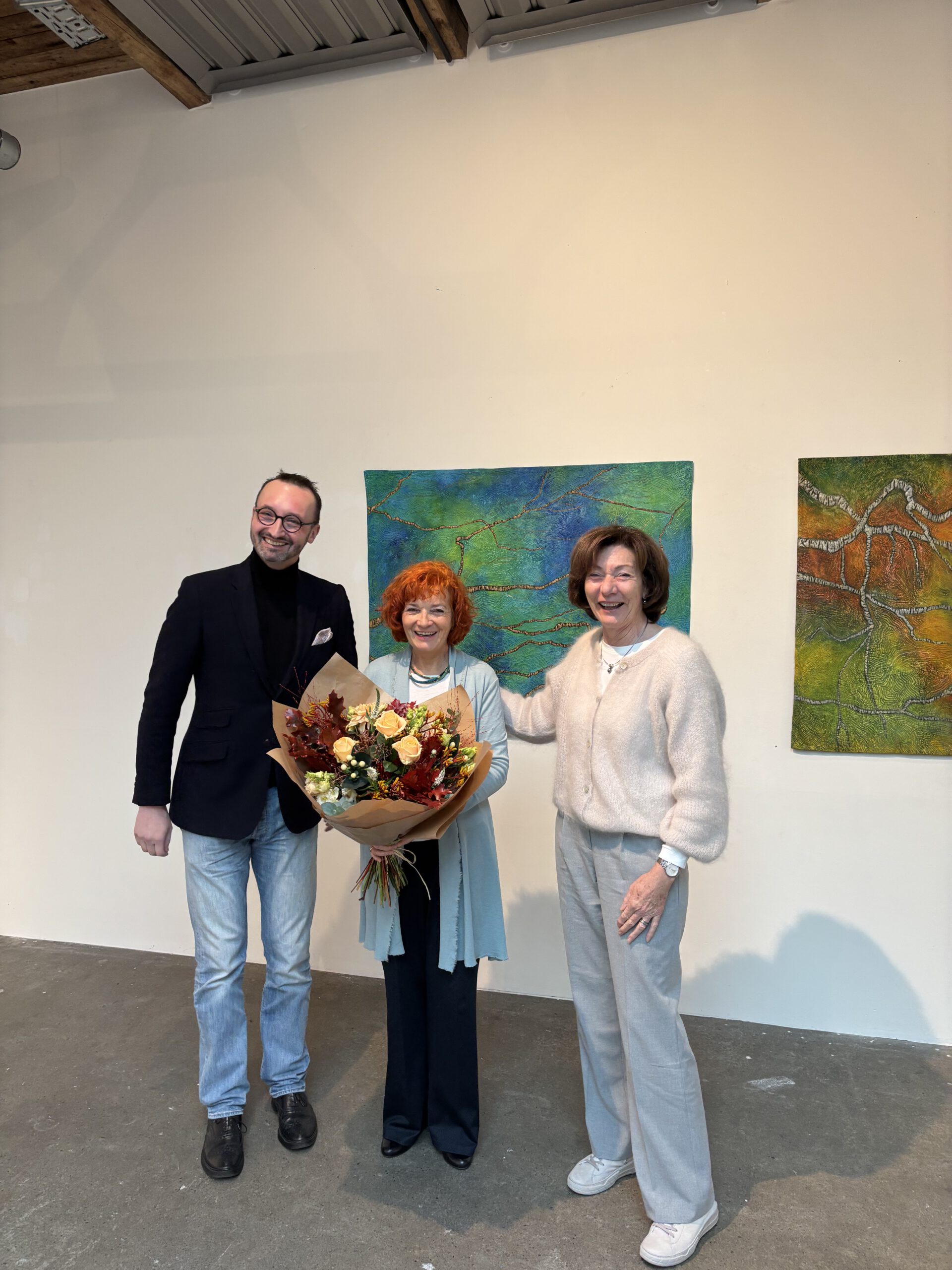 Drei Personen bei einer Ausstellungseröffnung mit Blumenstrauß.
