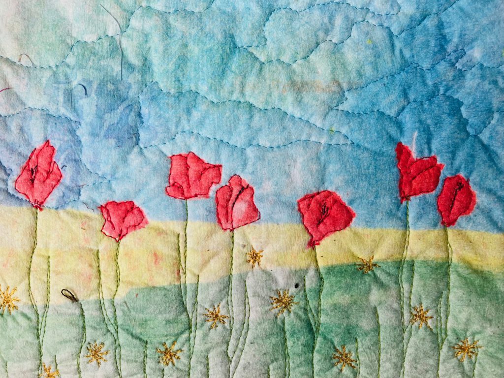 Gestickte rote Mohnblumen auf einem Hintergrund, der eine Landschaft mit blauem Himmel und grüner Wiese andeutet.